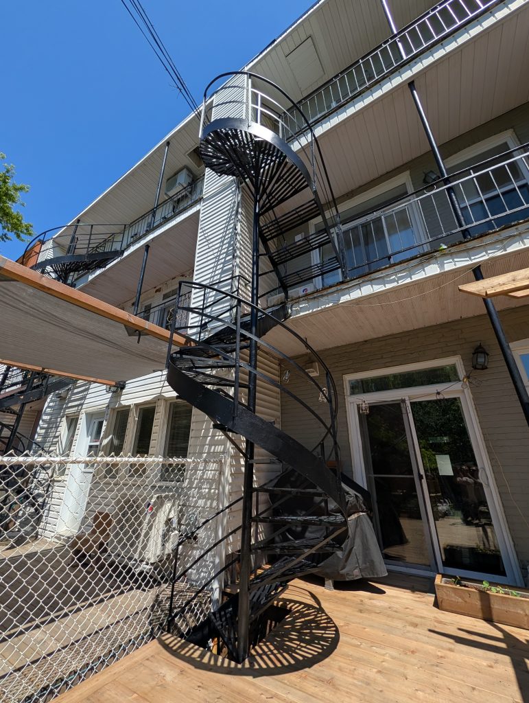 Black metal exterior spiral staircase attached to a multiunit building with balconies casting a circular shadow on a wooden deck  Bricopro  Bricopro