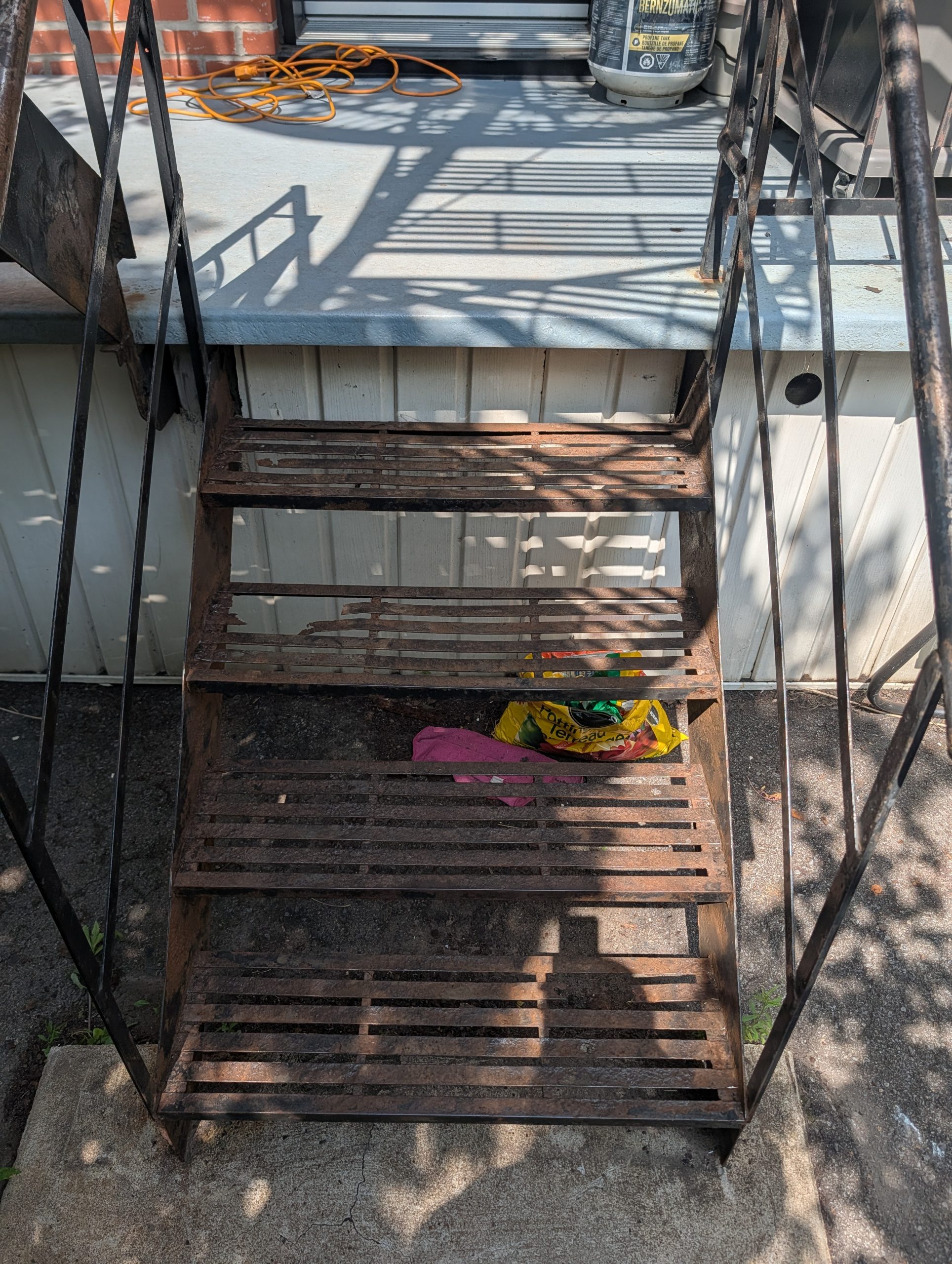 Outdoor rusted metal staircase descending to a concrete patio with shadows from the railing across the treads and a crumpled pink cloth and snack bag on the ground below  Bricopro