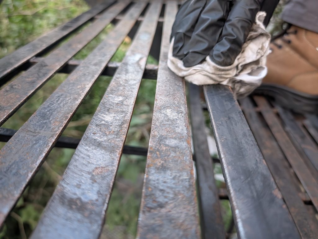 Gloved hand wiping a rusted metal bench slat outdoors with a cloth The bench slats appear weathered and rusted with greenery blurred in the background  Bricopro  Bricopro