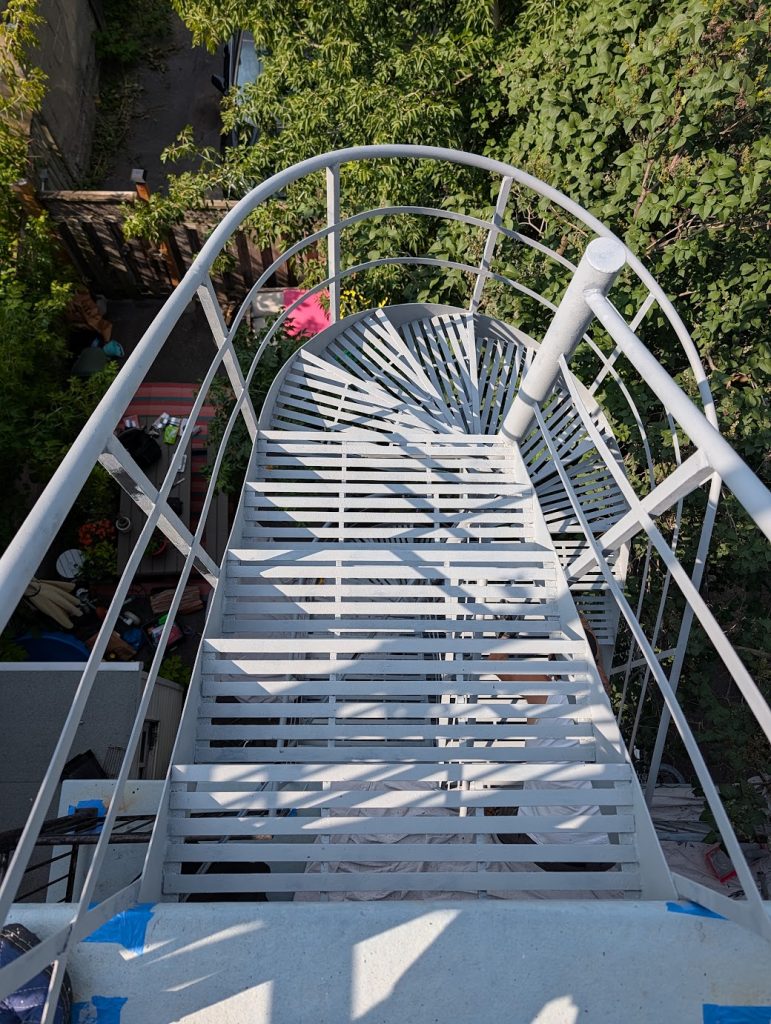Top down view of a grey metal spiral staircase with railings descending among green trees and a yard below  Bricopro  Bricopro